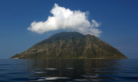 tiny rocky island with grassy upper slopes seen rising out of dark blue sea; the sky is also blue with a white fluffy cloud hovering over the island