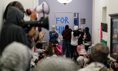 people in masks and keffiyehs sit and stand in a hall