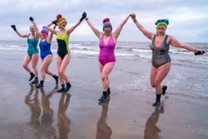 Swimmers in brightly coloured costumes and hats hold hands  on a beach