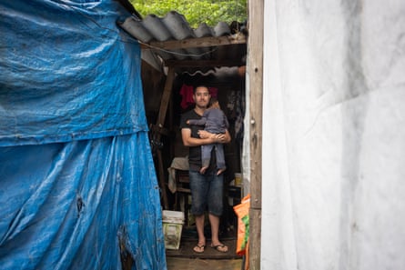 A man holding a small child stands under a corrugated roof in a makeshift house.