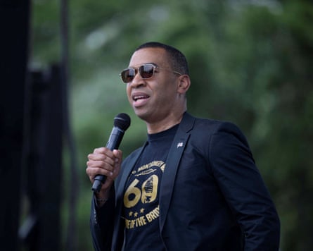 Man in sunglasses, T-shirt and blazer speaks into microphone.