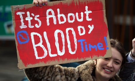 A young woman holding up a red-painted homemade sign saying 'It's about bloody time!'
