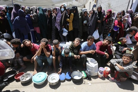 A group of children sit on the ground with food bowls and container with adults standing in line behind them