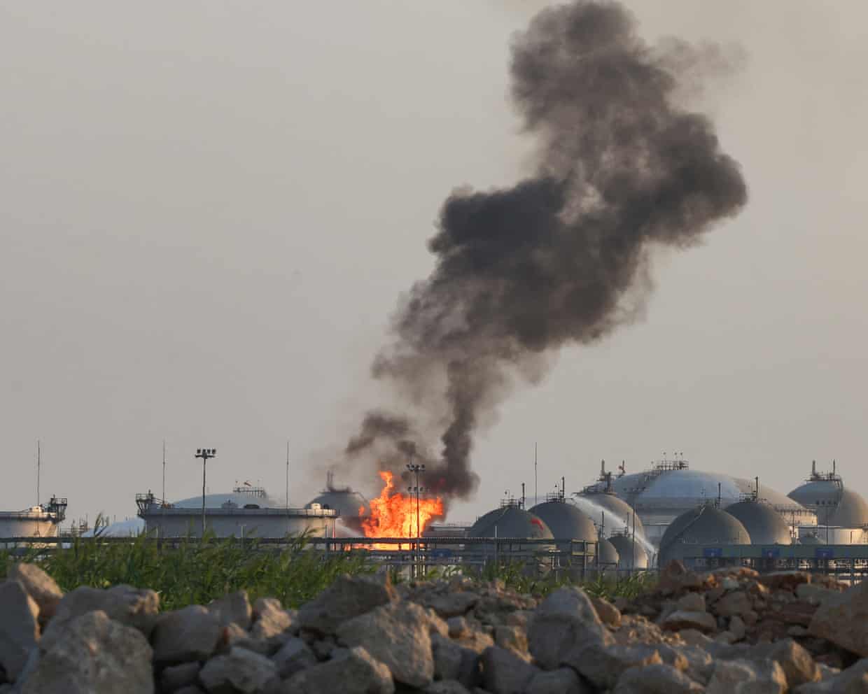 Flames and thick black smoke rise from Aramco’s Riyadh refinery, viewed over a rocky field