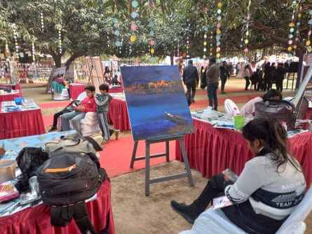Coloured discs hang in strings from trees, inder which various stalls have been set out. Closest to the camera is an easel with a large paining on it.