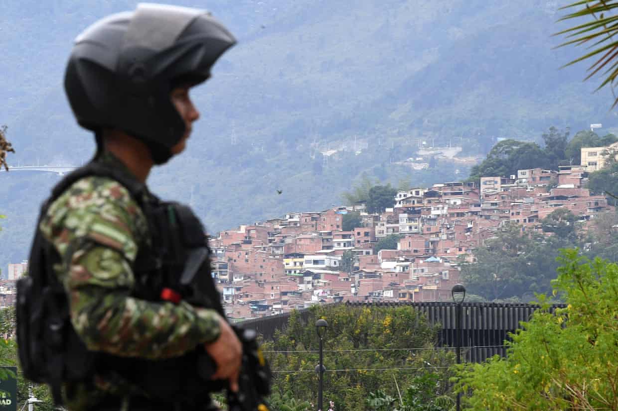 A soldier holding a gun stands guard, with houses clustered on a slope in the distance