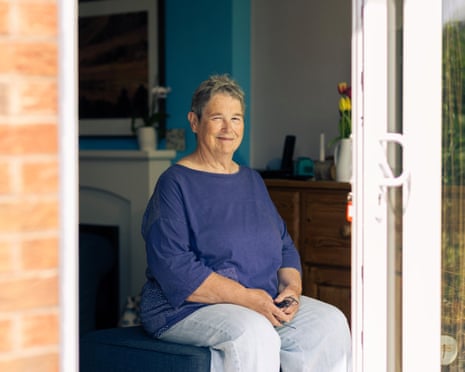 Maxine in purple top and jeans sits in her house, by open patio doors, smiling at the camera