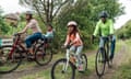 Family riding their bikes around a public park. The youngest girl is sitting in a support seat on the back of her mothers bike. They are wearing helmets and are social distancing.