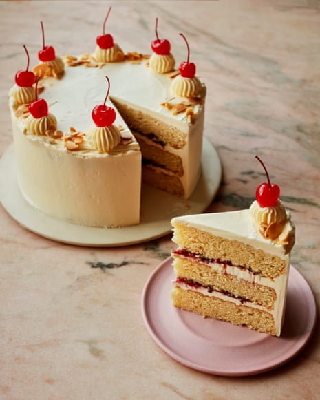 A cake with seven cocktail cherries on top sitting on a late on a marble countertop, and next to it a plate with a slice of cake showing its three layers and jam inside