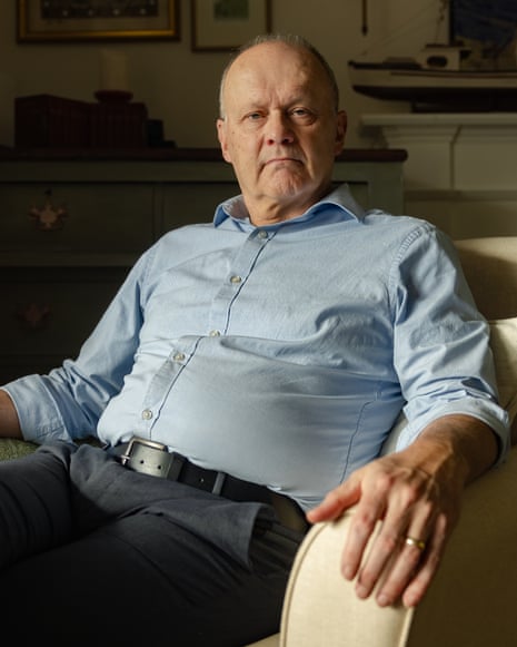 Chris Moore wearing a light-blue shirt and dark trousers, sitting in a cream armchair with his arms resting on the arms, looking at the camera
