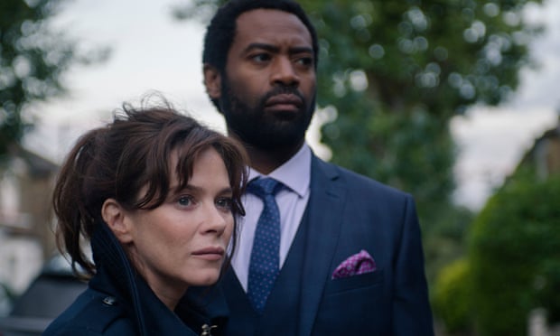 Friel wearing a dark blue raincoat looking serious standing next to a taller man in a suit