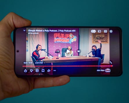 A hand holding a smartphone showing Meloni being interviewed on a podcast.