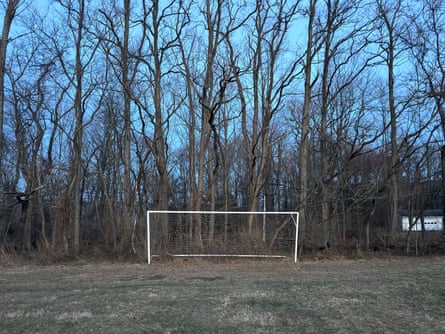 A soccer goal on a rough patch of grass with trees in the background.