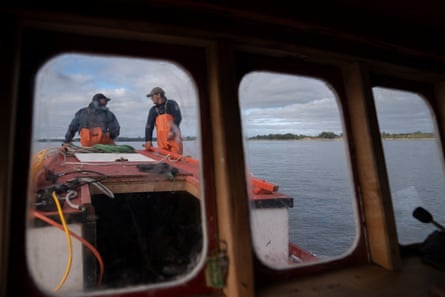 Two men on a boat wearing fishing gear can be seen through the boat’s window. The sea is behind them