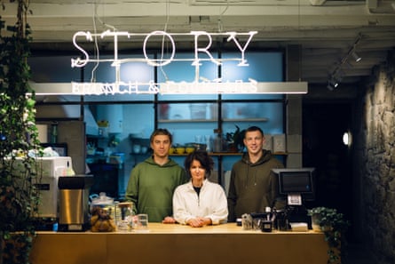 A woman stands at a cafe counter with two men with a neon sign above their heads saying ‘Story’