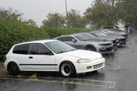 A white 1990s Honda Civic parked in rain ahead of newer sports cars at a car show