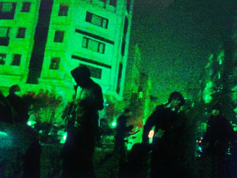 Very grainy image of a street scene at night in Iran during protests, bathed in green light, people milling around in hoods and with faces covered, no one identifiable.