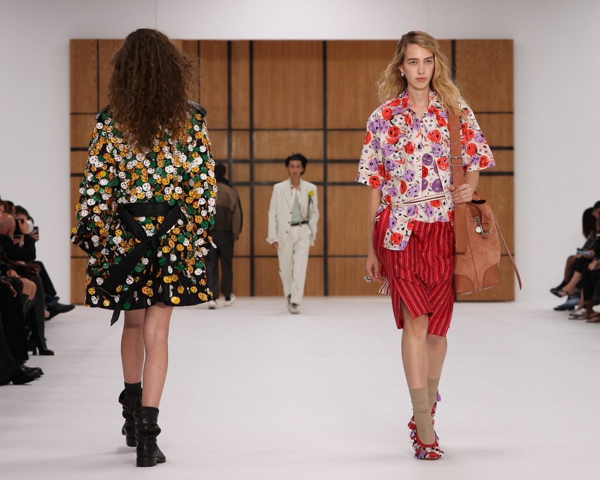 A model wearing green, white and yellow smiley-face buttons on a thigh-length coat passes another model wearing a red, purple and white smiley faces on a blouse and a red-and-black-striped skirt