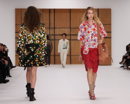A model wearing green, white and yellow smiley-face buttons on a thigh-length coat passes another model wearing a red, purple and white smiley faces on a blouse and a red-and-black-striped skirt