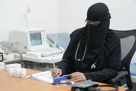 A woman in a black burqa makes notes. She has a stethoscope around her neck and is surrounded by medical equipment