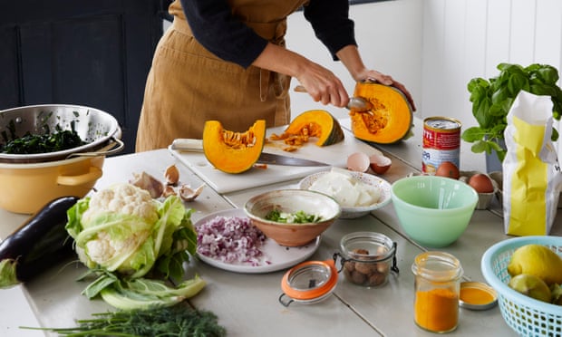 A woman in an apron scoops pumpkin seeds out of a cut pumpkin. In front of her on the bench are an array of ingredients including cauliflower, onions, canned tomatoes and spices