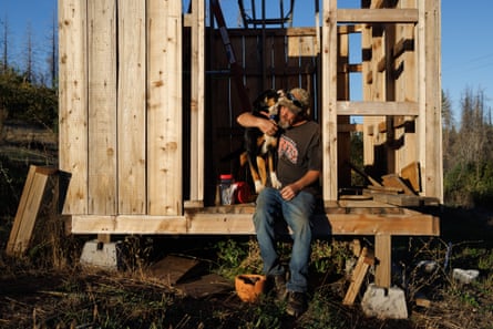An older white man sits on the raised wood foundation, his arm around a dog, in bright light from the setting sun.