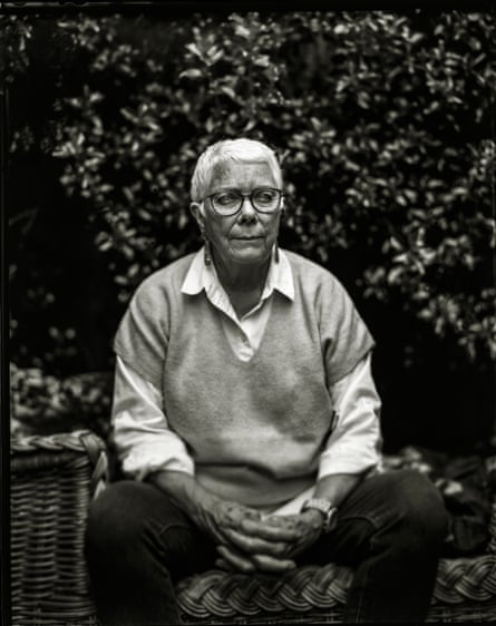A woman with short hair and glasses sits with her hands clasped in a garden in a black and white image