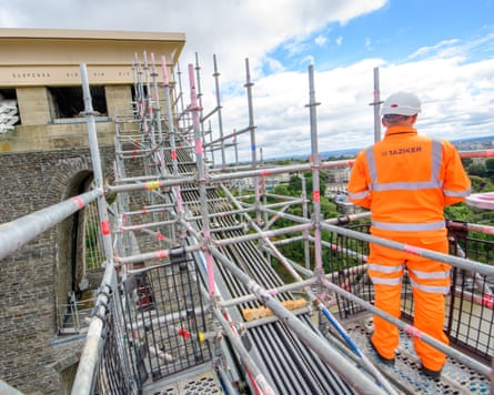 A worker in hi-vis clothing an white hat stands on a raise platform during refurbishment of the chain links on Clifton Suspension Bridge.