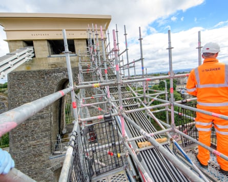 A man in orange hi-vis overalls and white helmet stands on scaffolding around the stone tower, which has the motto engraved at its top