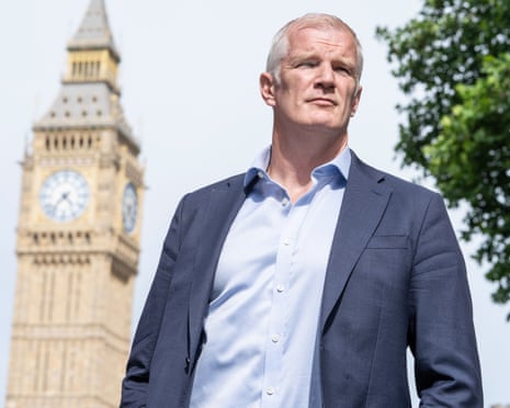 Carns in a grey suit jacket and blue shirt stands outdoors with Big Ben in the background