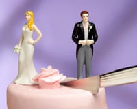 A knife cuts between bride and groom figurines standing on opposite sides of a pink wedding cake
