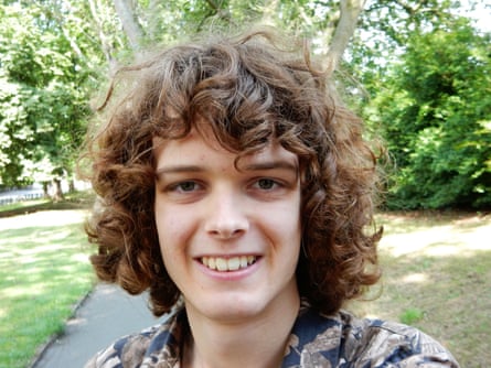 he is 19 with shoulder-length brown curly hair, and is smiling as he stands in a park.