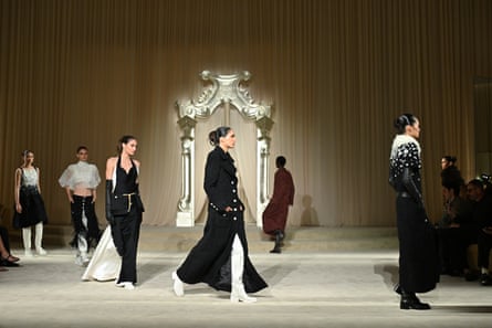 Models walk on pale cream carpet with pale curtains and a silver archway in the background; they are wearing long black coats, skirts and loose trousers and some have thigh length white boots with chunky heels