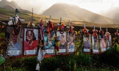 Long row of laminated color photos of people, attached to a fence with white crosses and colorful ribbons, with sun peeking through clouds beyond mountain behind it.