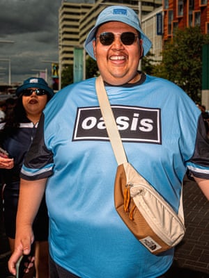 A man wearing an Oasis T-shirt and round sunglasses grins.