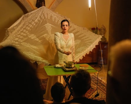 A woman in a long lace dress stands in front of a table with a pan on a burner with a lace backdrop behind her