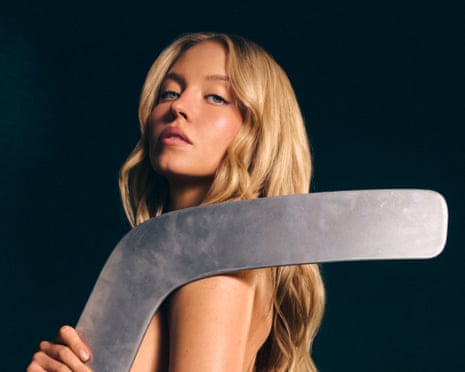 Sydney Sweeney rests a large metal hockey stick blade on her bare shoulder
