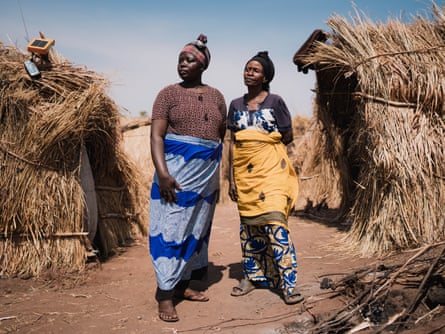 Two women stand between straw-covered huts.