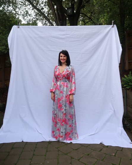 A woman in a floral pink long-sleeved dress stands in front of a white fabric backdrop in a leafy courtyard