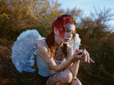 A young woman covered in tattoos and wearing a vest and angel wings looks forlorn with evergreen trees in the background