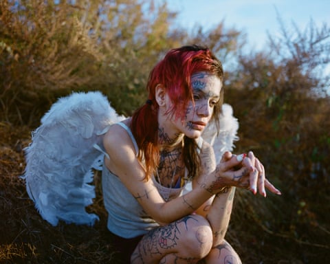 A woman with tattooed face and body, wearing angel wings, crouches outdoors looking down