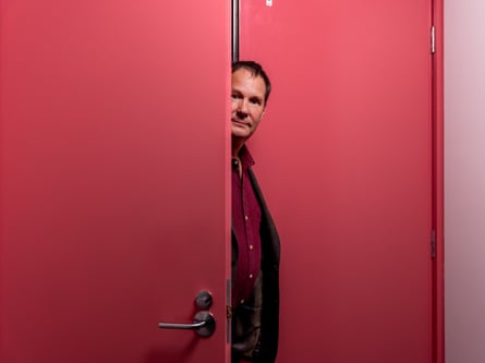 A man in a brown suit walks through a red door he is opening
