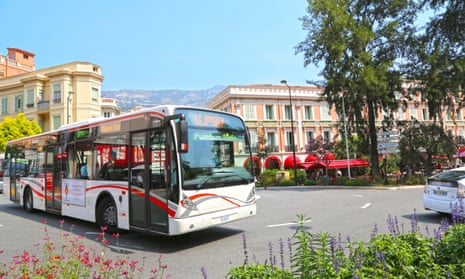 A bus in Monaco with hills and stuccoed buildings behind, flowers in the foreground.
