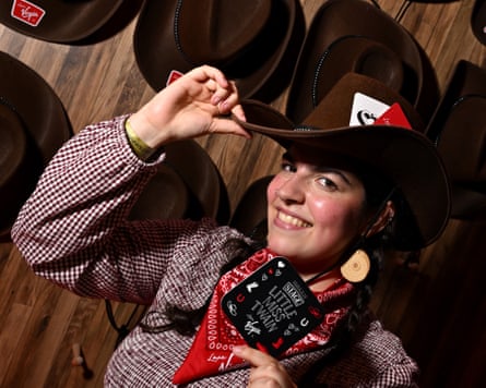 A woman touches the peak of her cowboy hat in front of a wall of hats.