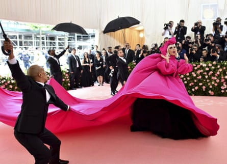 Lady Gaga in 2021 wear a flowing pink gown and surrounded by people in tuxedos with black umbrellas