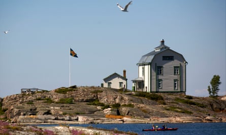 Two people in a red canoe row past a traditional white timber-built house situated on the water’s edge