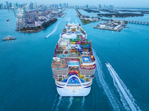 A huge ship, with hundreds of crew members seen as tiny figures on the many decks, approaching a US city