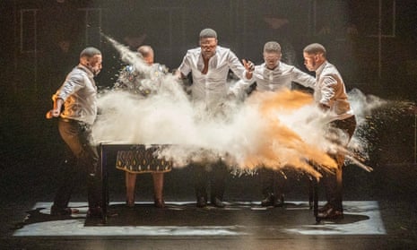 Five dancers pound a table covered in dust, causing it to fly up