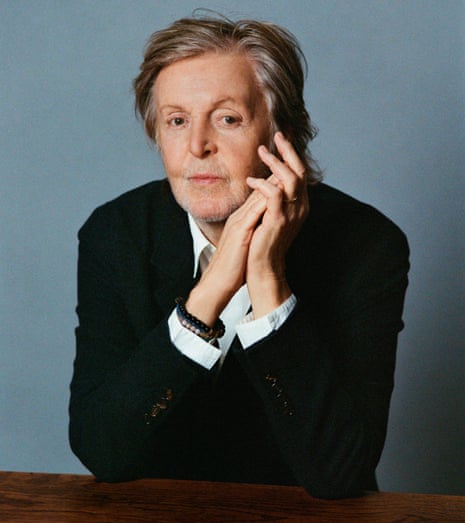 A man wearing a dark blazer, a white shirt, beaded bangles, with his hands together