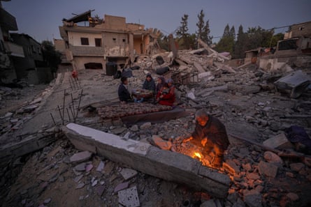 A family sit on a rug amid rubble as a man tends to a fire